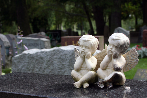 Kaksi kivienkeliä istuu jalat ristissä, kädet poskilla hautakiven päällä.