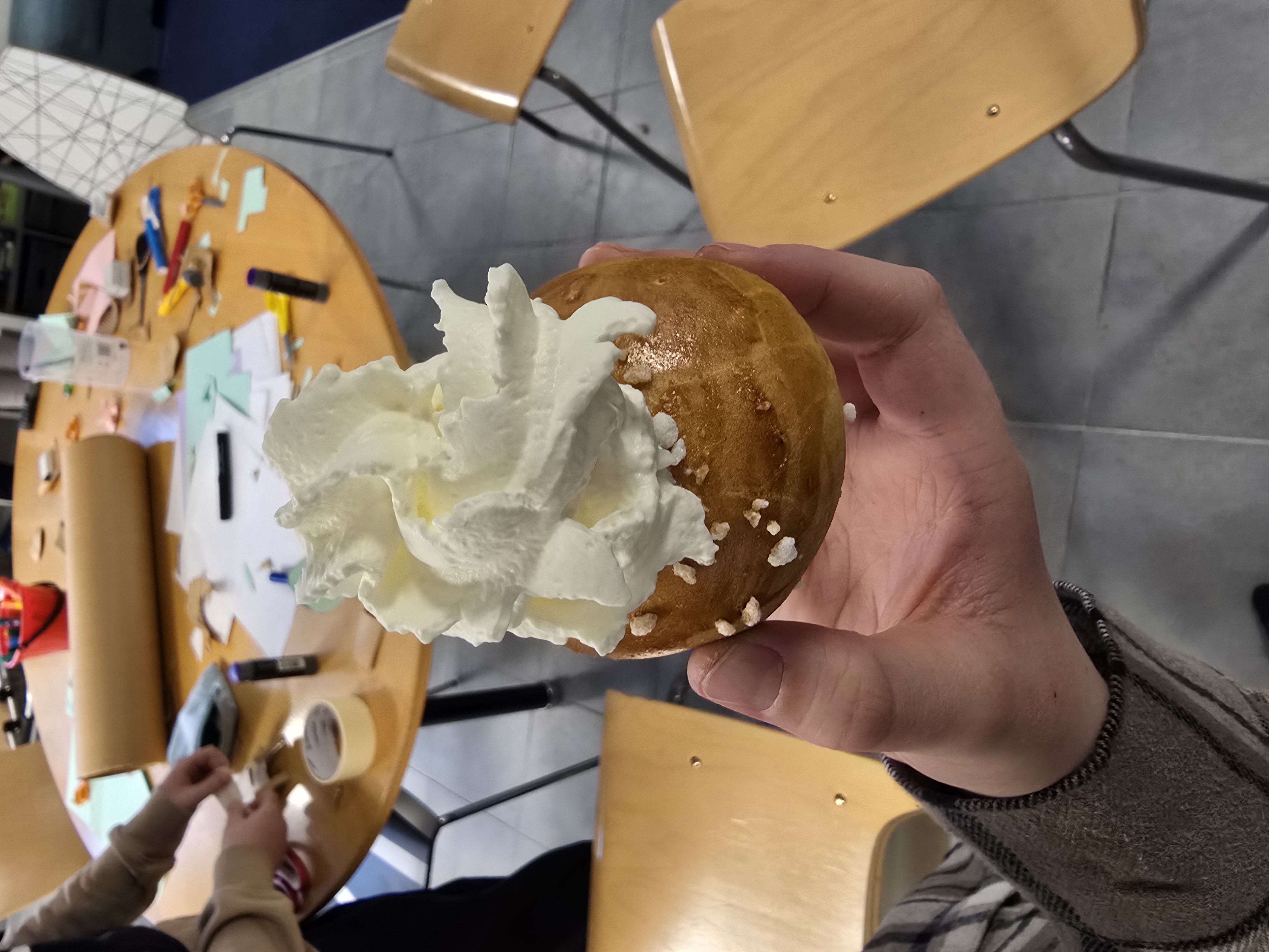Kuvassa on henkilön kädessä pulla, jonka päällä on iso keko kermavaahtoa.